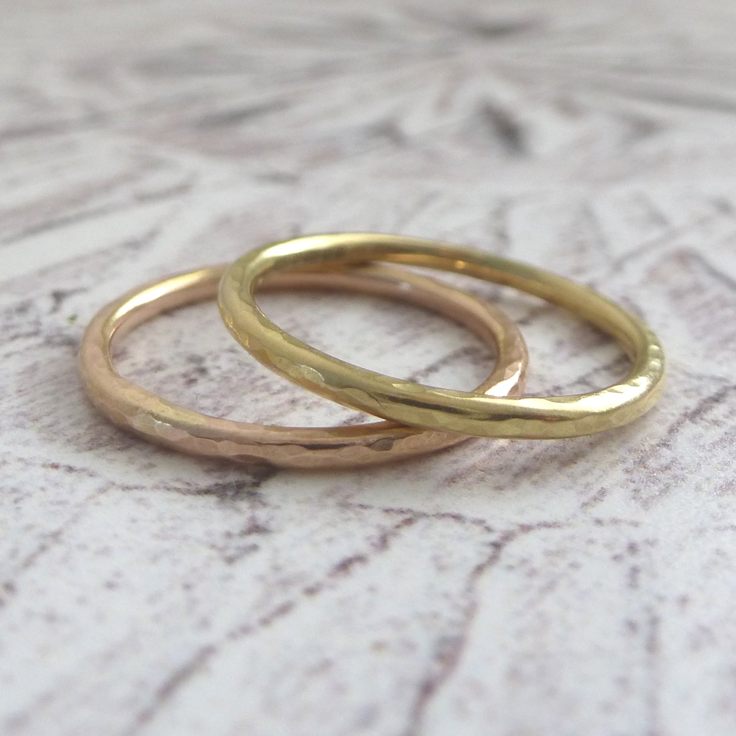 A pair of 18ct yellow gold slim band, one yellow one rose, both with hammered finish. Close up sitting on a board.