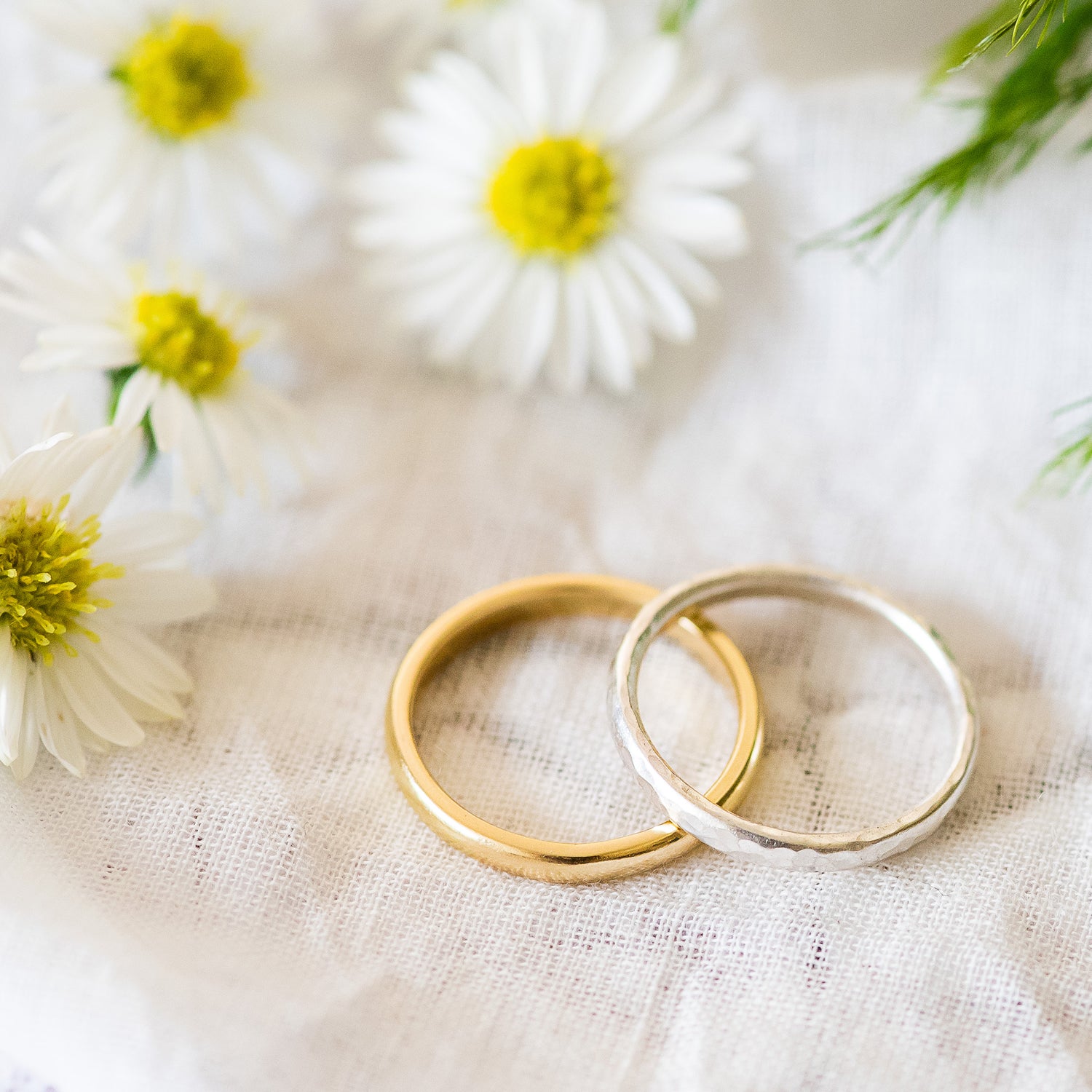 A pair of slim D shaped wedding bands, one 18ct yellow gold one sterling silver, sat on fabric, with some flowers