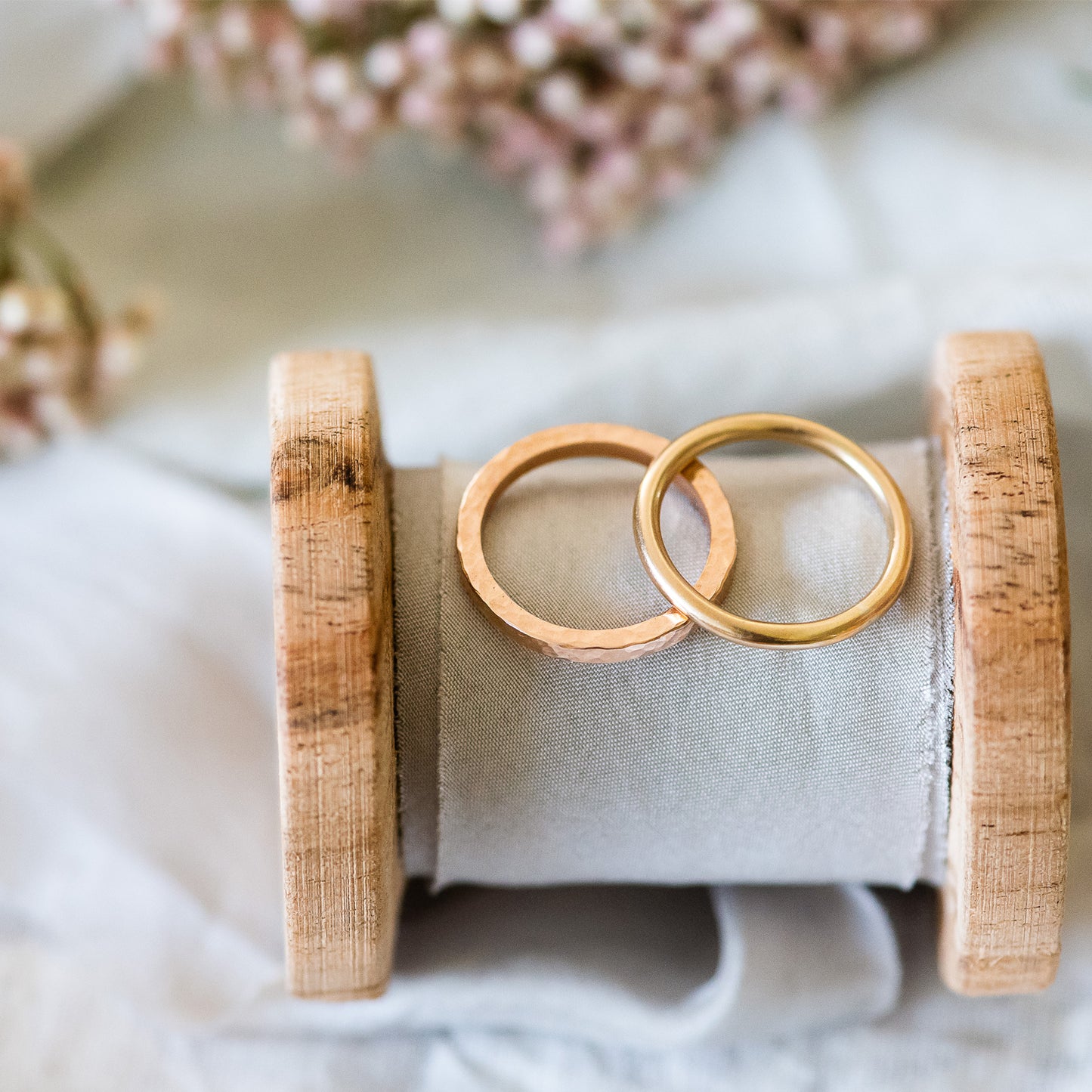 A pair of 9ct gold wedding rings, one hammered square and one smooth round, sitting on a ribbon wound around a wooden bobbin