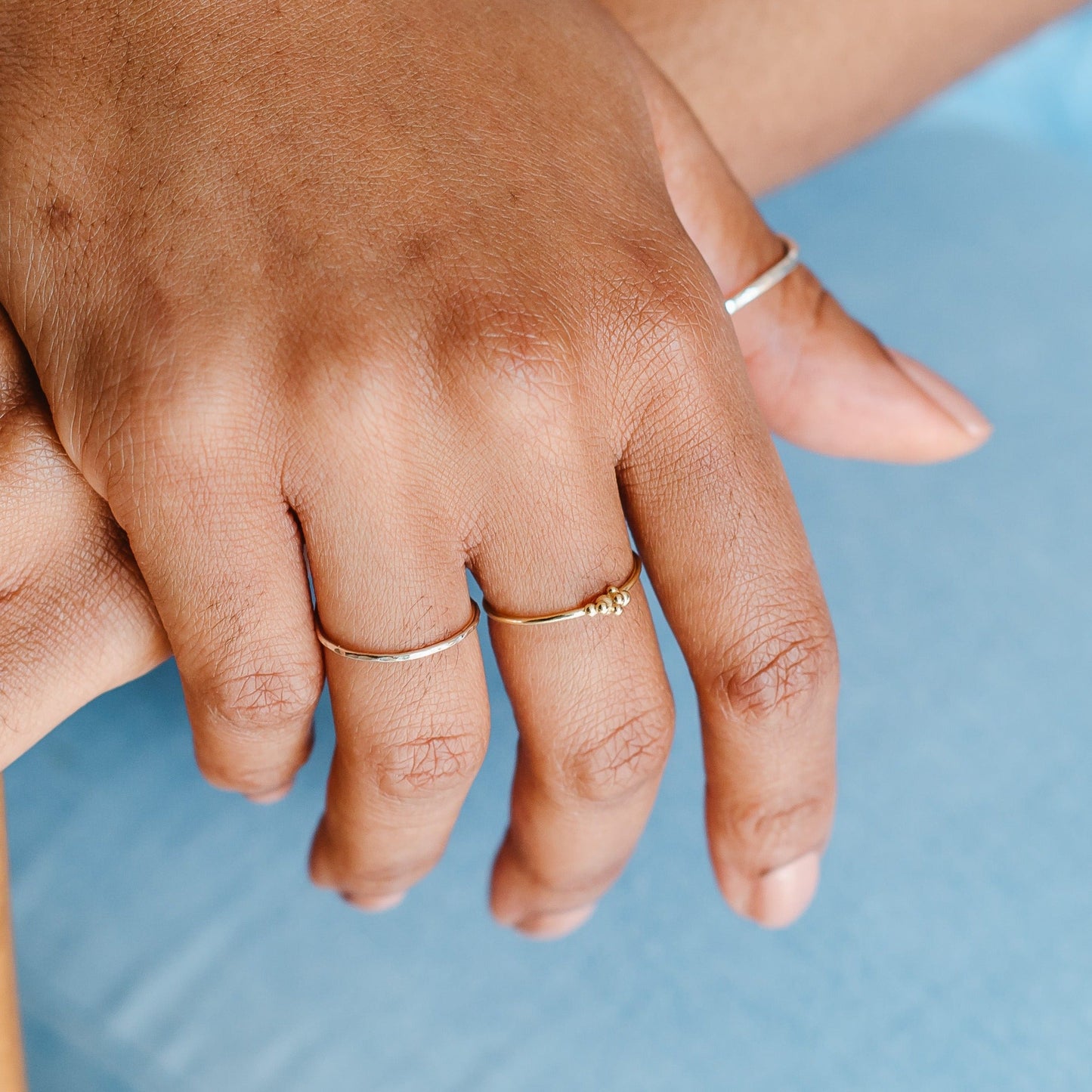 A gold bubble ring and a thin hammered band being worn, blue background behind the hand.