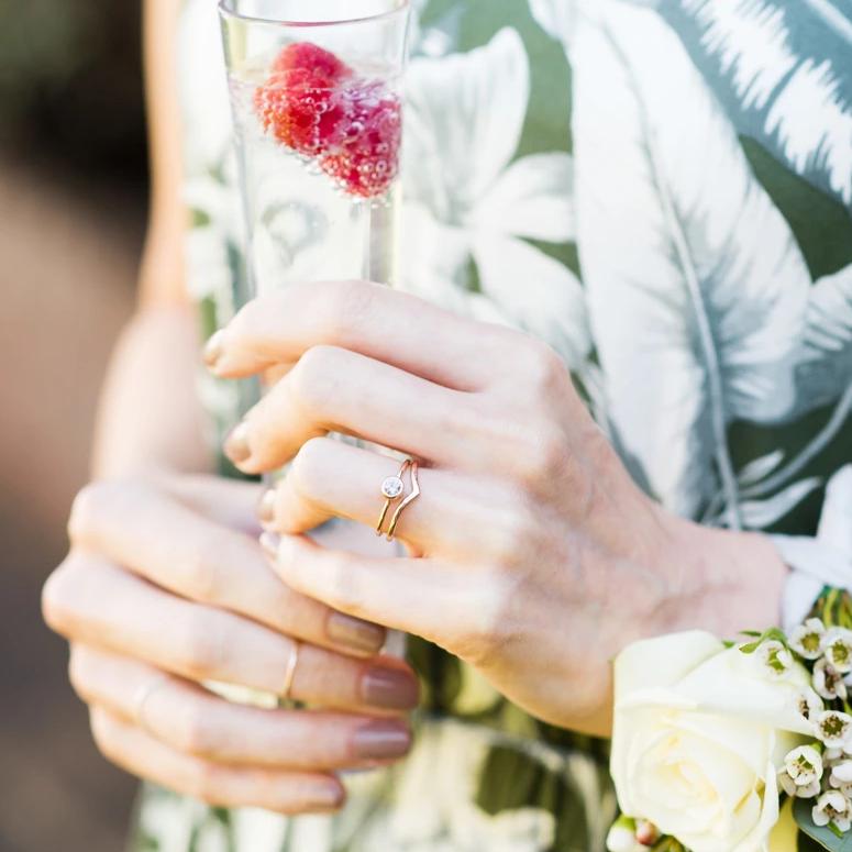 A set of two rings, one spinel ring and one 9ct rose gold wishbone, being worn. The hand is holding a flute. 