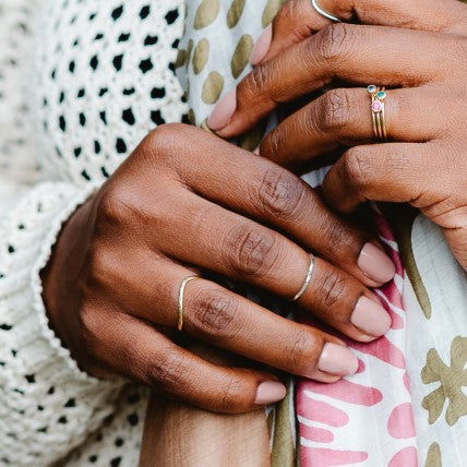 Two hands holding a scarf, wearing a selection of rings, a hammered curved band and some stone set stacking rings. 
