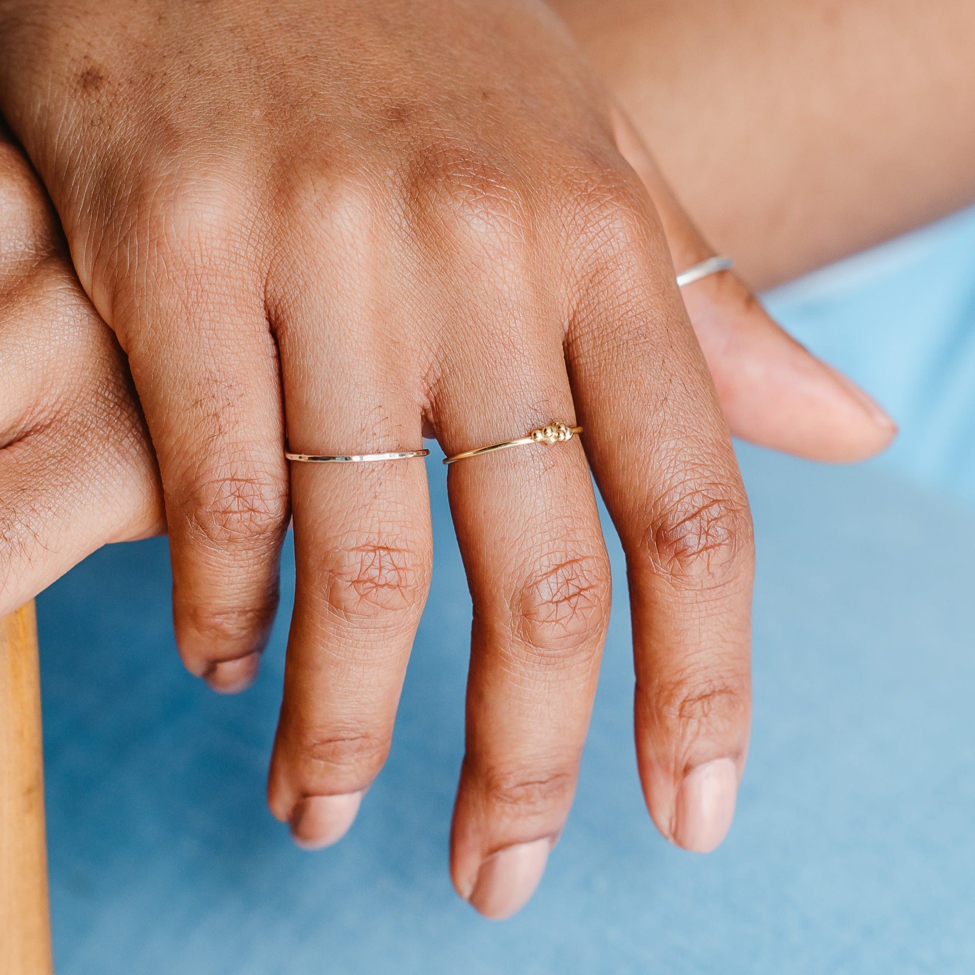 A hand with several skinny rings, one bubble ring and one hammered band in front of a blue background.