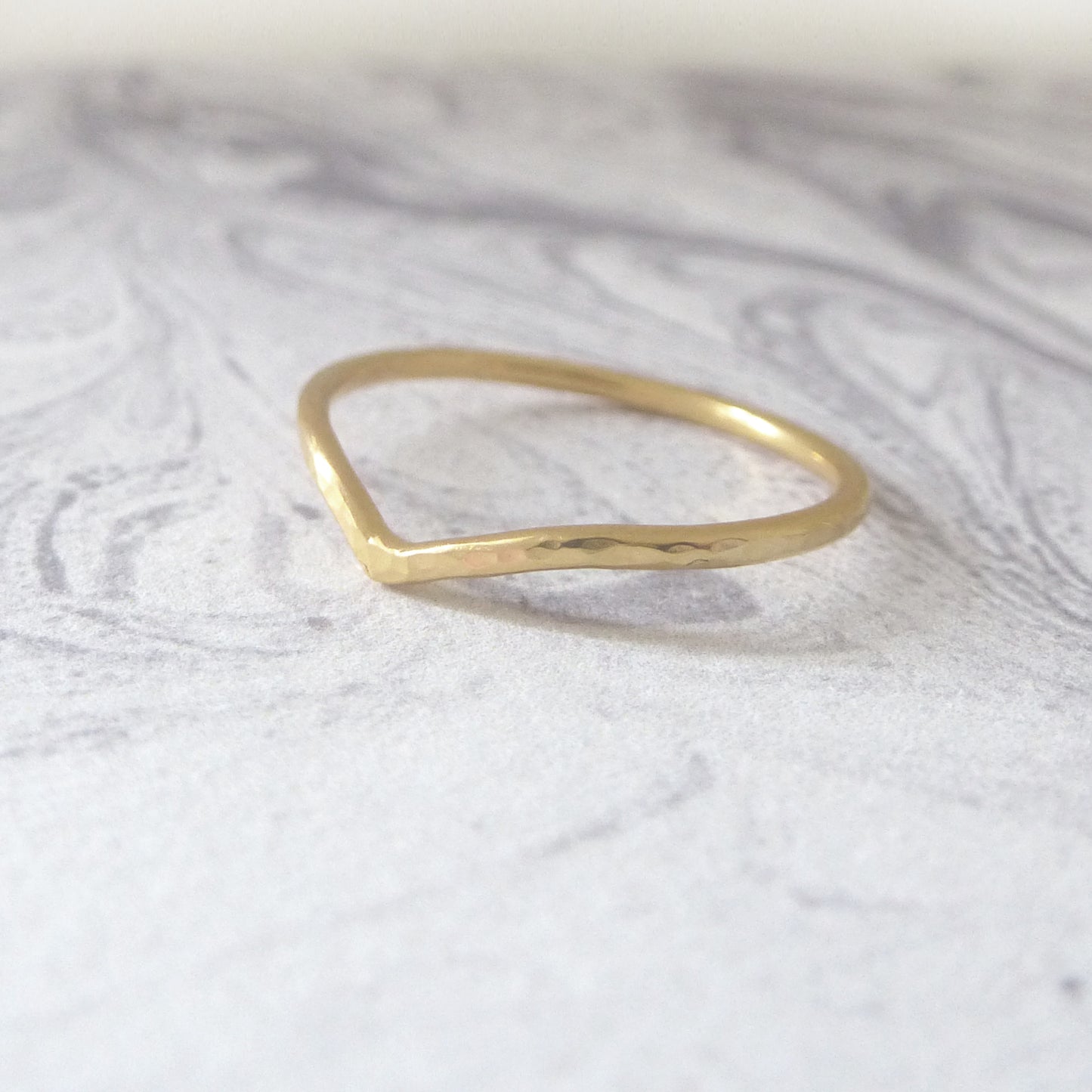 Thin wishbone wedding ring in 18ct yellow gold, hammered finish, sitting on a marbled background.