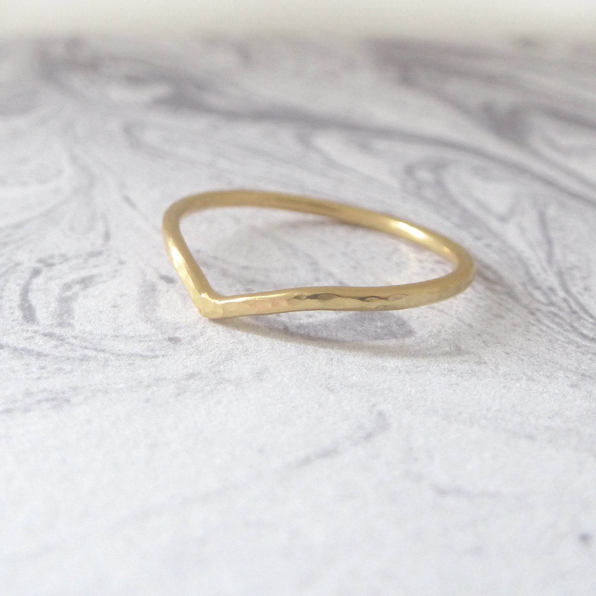 Thin wishbone wedding ring in 18ct yellow gold, hammered finish, sitting on a marbled background.