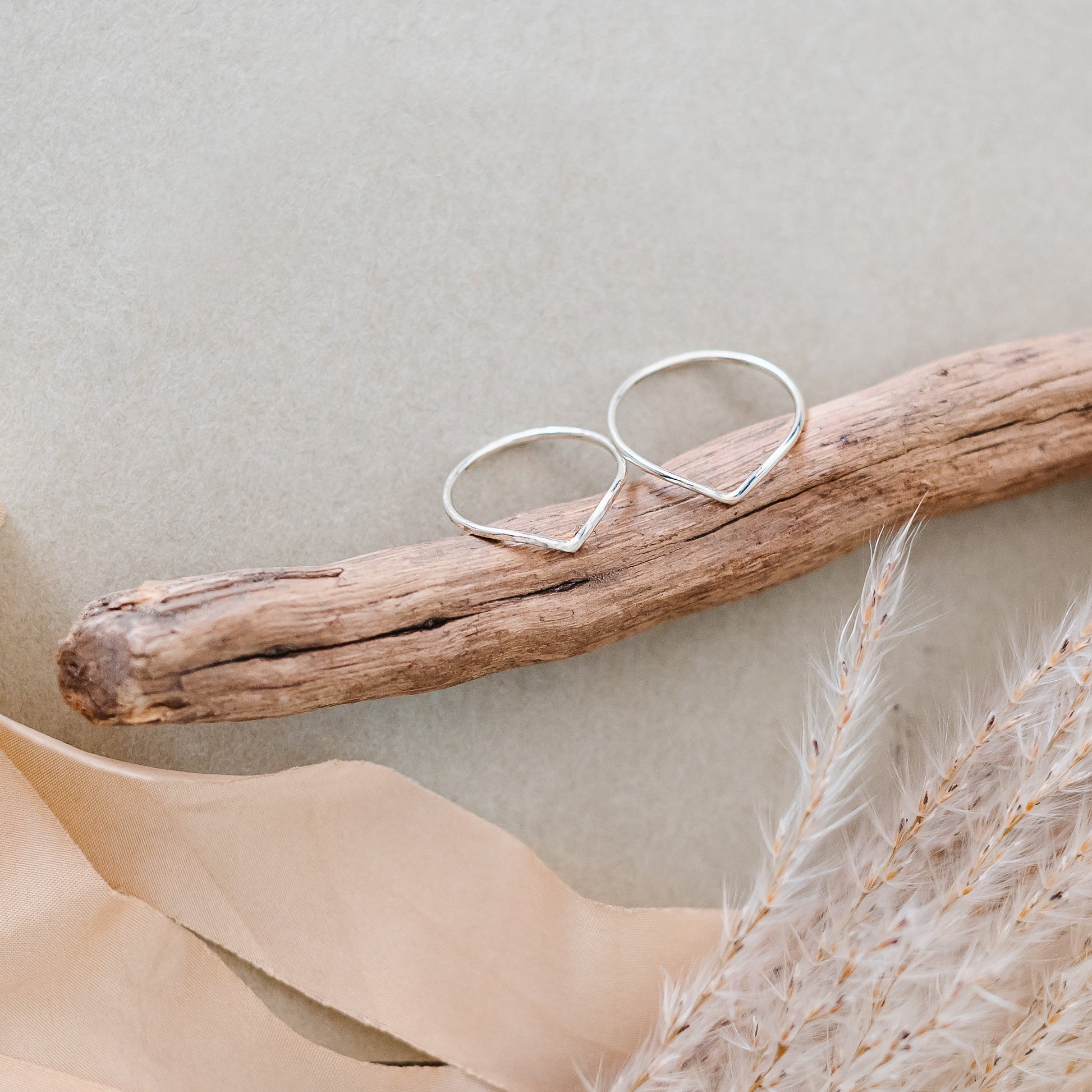 Two slim wishbones in 9ct white gold, one hammered one smooth. Wishbones are leaning on a piece of wood, with dried flowers and ribbon in the background