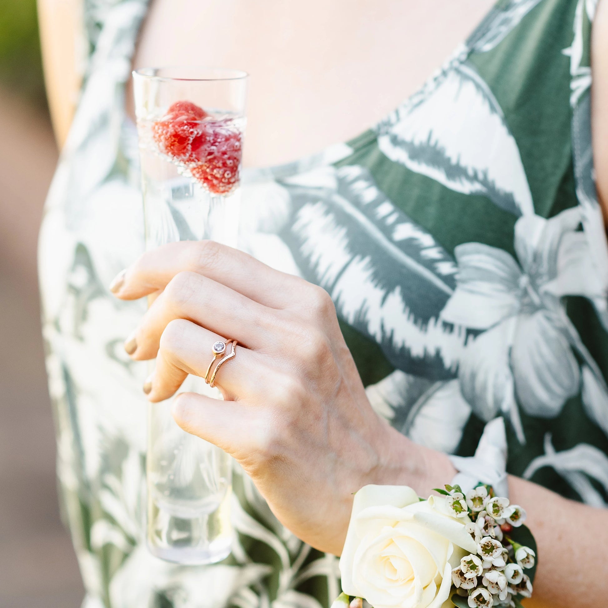 A spinel and 9ct rose gold ring, with 9ct rose gold wishbone ring, being worn. The hand is holding a champagne flute and wearing a corsage.